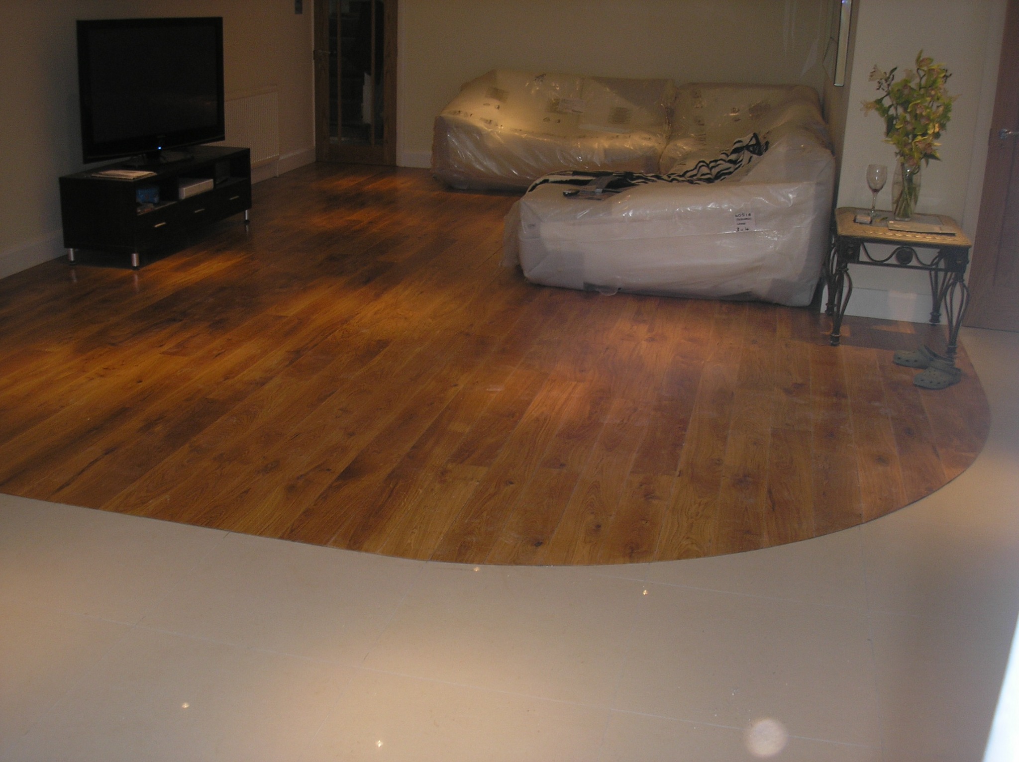 Połączenie drewnianej podłogi z jasnymi, kwadratowymi płytkami podłogowymi, na tle meble owinięte folią ochronną i telewizor na szafce.