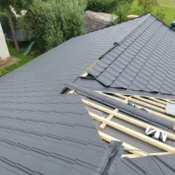 Fragment dachu w trakcie układania, widoczna konstrukcja drewniana, membrana dachowa, częściowo ułożone szare panele dachowe i narzędzia. W tle budynek mieszkalny, trawnik i drzewa.