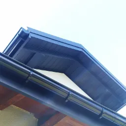 Niebieska rynna dachowa zamontowana na drewnianej konstrukcji, widok z dołu na tle jasnego nieba.