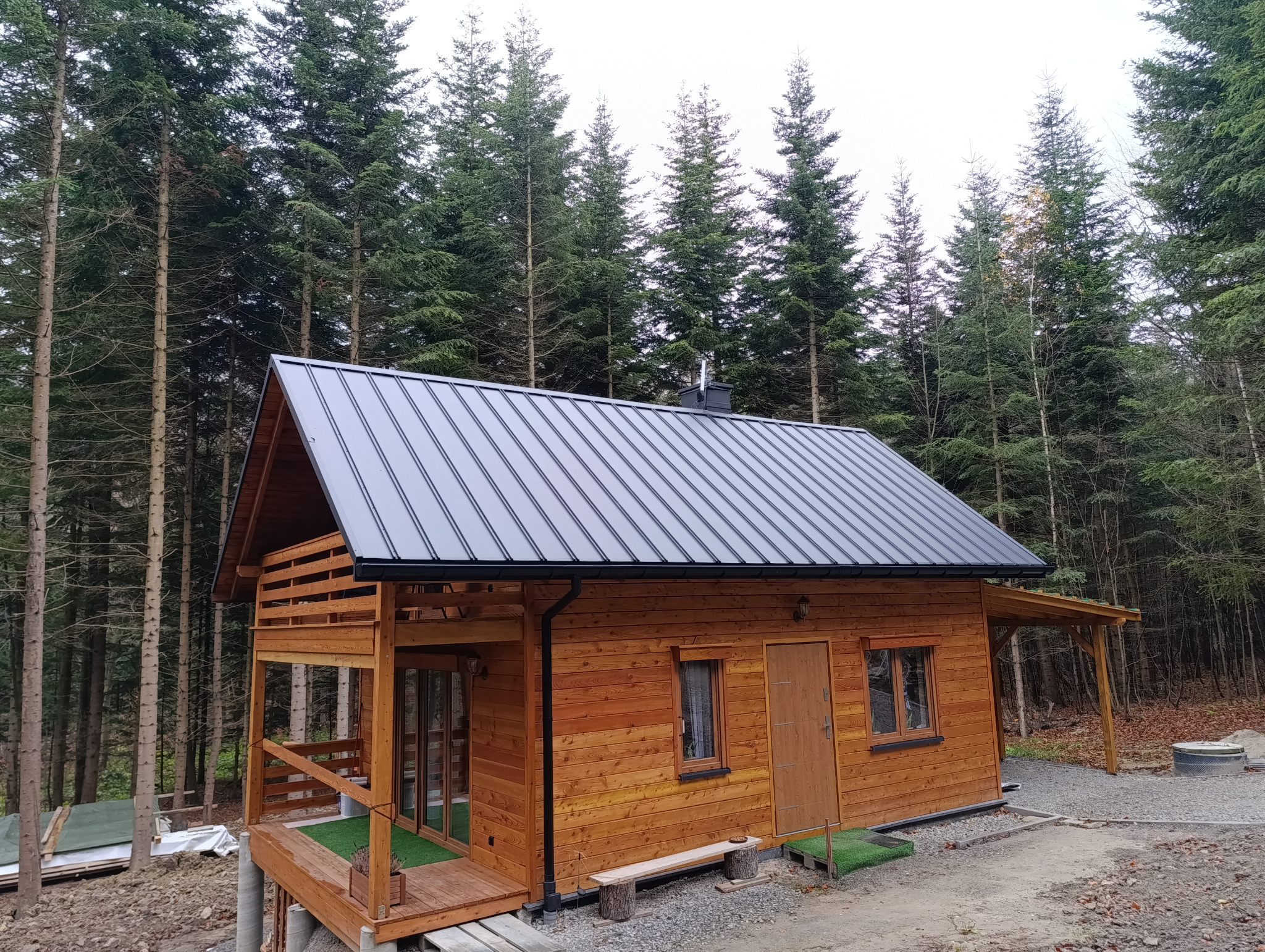 Drewniany domek letniskowy z nowym dachem z blachy trapezowej w kolorze grafitowym, na tle lasu iglastego.