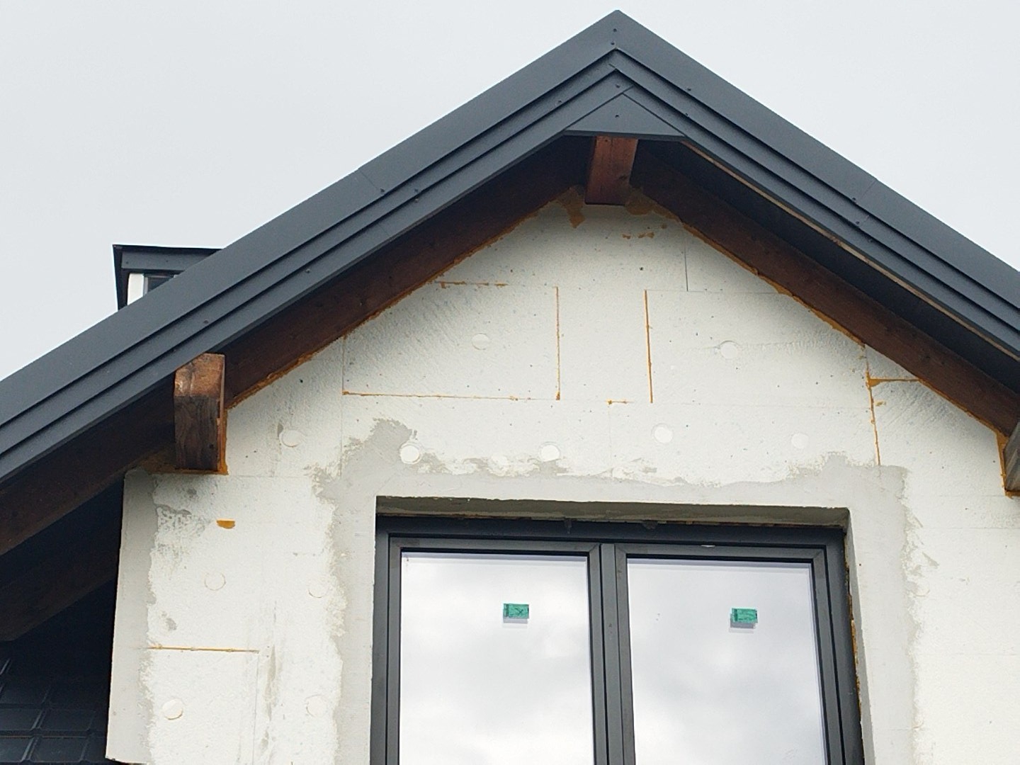 Dom w trakcie ocieplania dachu, widoczne szare obróbki blacharskie, drewniana więźba dachowa i biały styropian, okno z naklejkami.