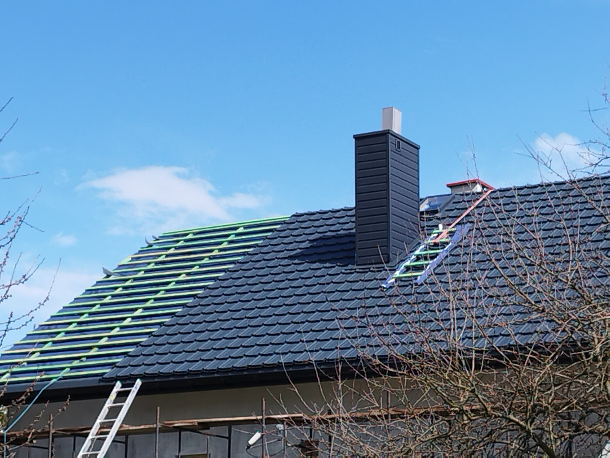 Częściowo pokryty ciemnoszarą dachówką dach domu z kominem, widoczne łaty dachowe w kolorze zielonym i drabina przy ścianie, błękitne niebo w tle.