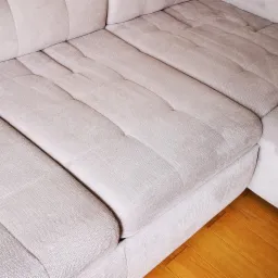 Jasna, narożna kanapa z widocznymi śladami czyszczenia tapicerki, ukazująca różnicę w odcieniu między wyczyszczoną a nieoczyszczoną powierzchnią, z fragmentem parkietu w dolnym rogu.