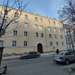 Odnowiona elewacja kamienicy z kremowym tynkiem, widoczna z poziomu ulicy, z zaparkowanymi samochodami i drzewami bez liści.