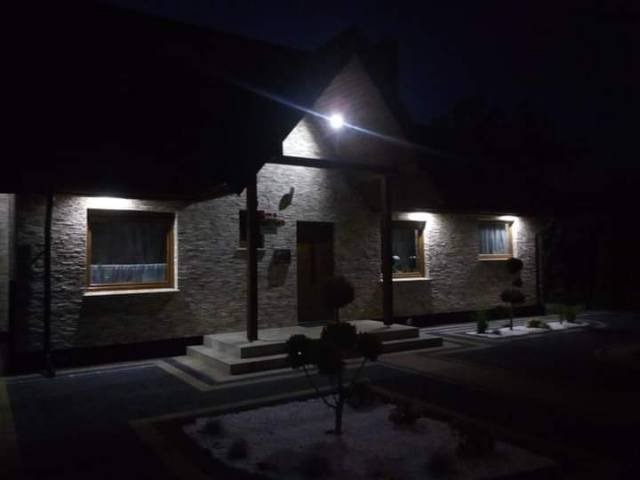 Elewacja domu jednorodzinnego z kamienną okładziną, oświetlona nocą reflektorem i lampami nad oknami, widoczne schody wejściowe i fragment ogrodu.