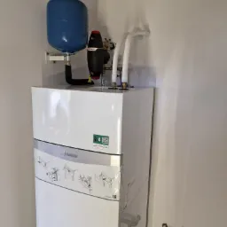 Biały kondensacyjny kocioł gazowy Vaillant z niebieskim zbiornikiem wyrównawczym i czarnym naczyniem przeponowym Flamco, zainstalowany na białej ścianie w pomieszczeniu w trakcie wykańczania.