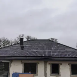 Dom w stanie surowym z panelami słonecznymi na dachu pokrytym ciemną dachówką, widok na szare niebo i drzewa bez liści w tle.