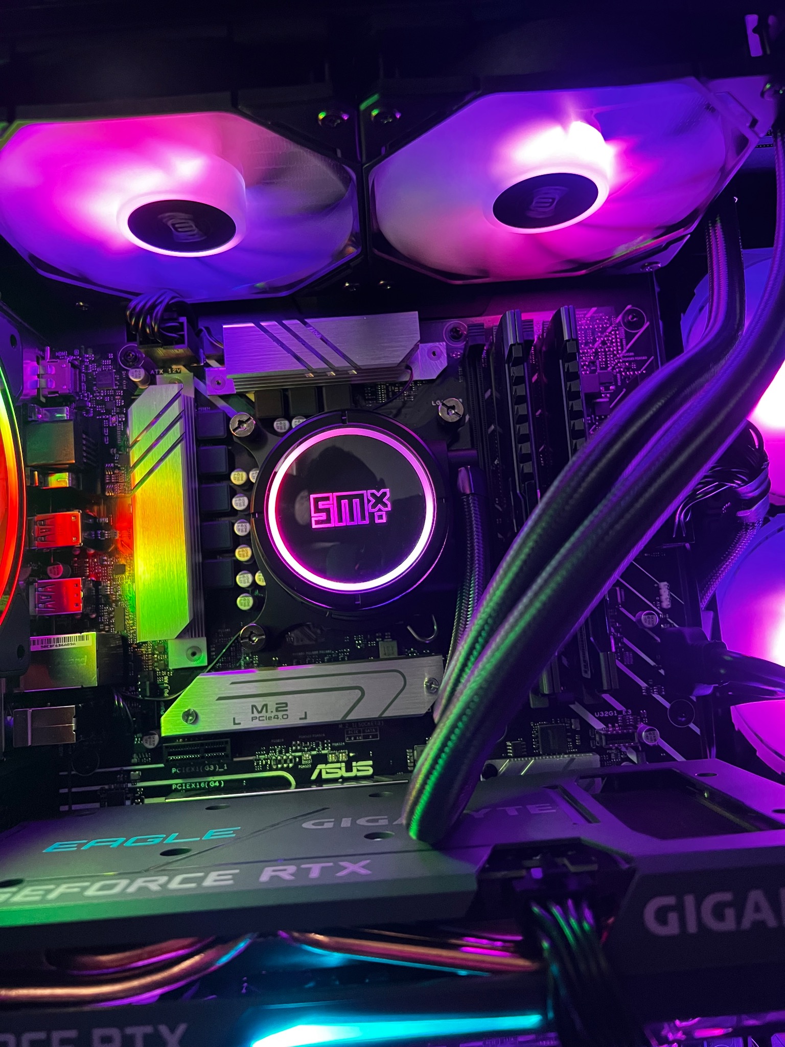 Wnętrze komputera PC z podświetlanymi wentylatorami chłodzenia procesora i karty graficznej RTX, widoczna płyta główna ASUS, chłodzenie wodne z logo SMX, pamięć RAM i złącza PCI Express.