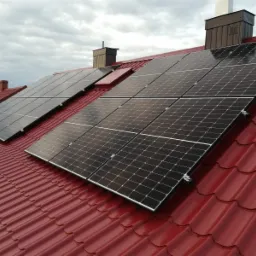 Panele fotowoltaiczne zamontowane na czerwonym dachu z blachodachówki w Sołonce, widoczne kominy wentylacyjne i pochmurne niebo.