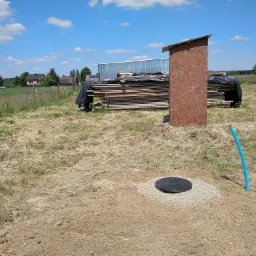 Studnia kręgowa z czarną pokrywą w trakcie budowy, widoczna niebieska rura doprowadzająca wodę, w tle stos drewna i budynek gospodarczy.