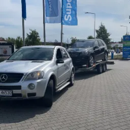 Srebrny samochód osobowy marki Mercedes holuje na przyczepie inny, czarny samochód Mercedes na tle stacji benzynowej i flag reklamowych.