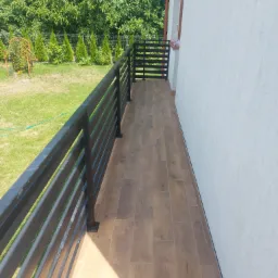 Wyłożony płytkami taras z czarną metalową balustradą, widok na trawnik i żywopłot z iglaków w słoneczny dzień.