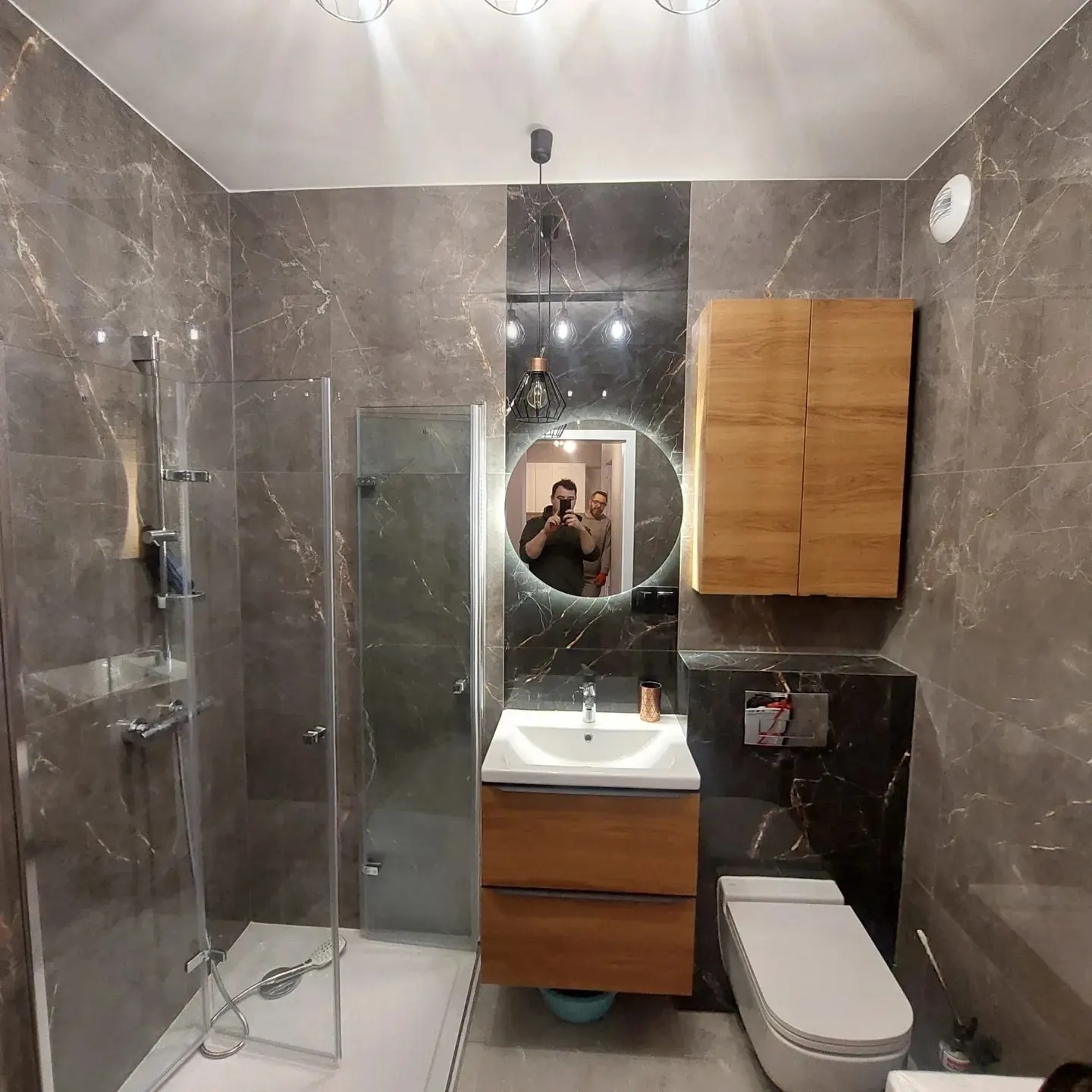 Wykończona łazienka z szarymi marmurkowymi płytkami, kabiną prysznicową ze szklanymi drzwiami, podświetlanym okrągłym lustrem, drewnianą szafką pod umywalką i wiszącą szafką na ścianie, z widocznym...