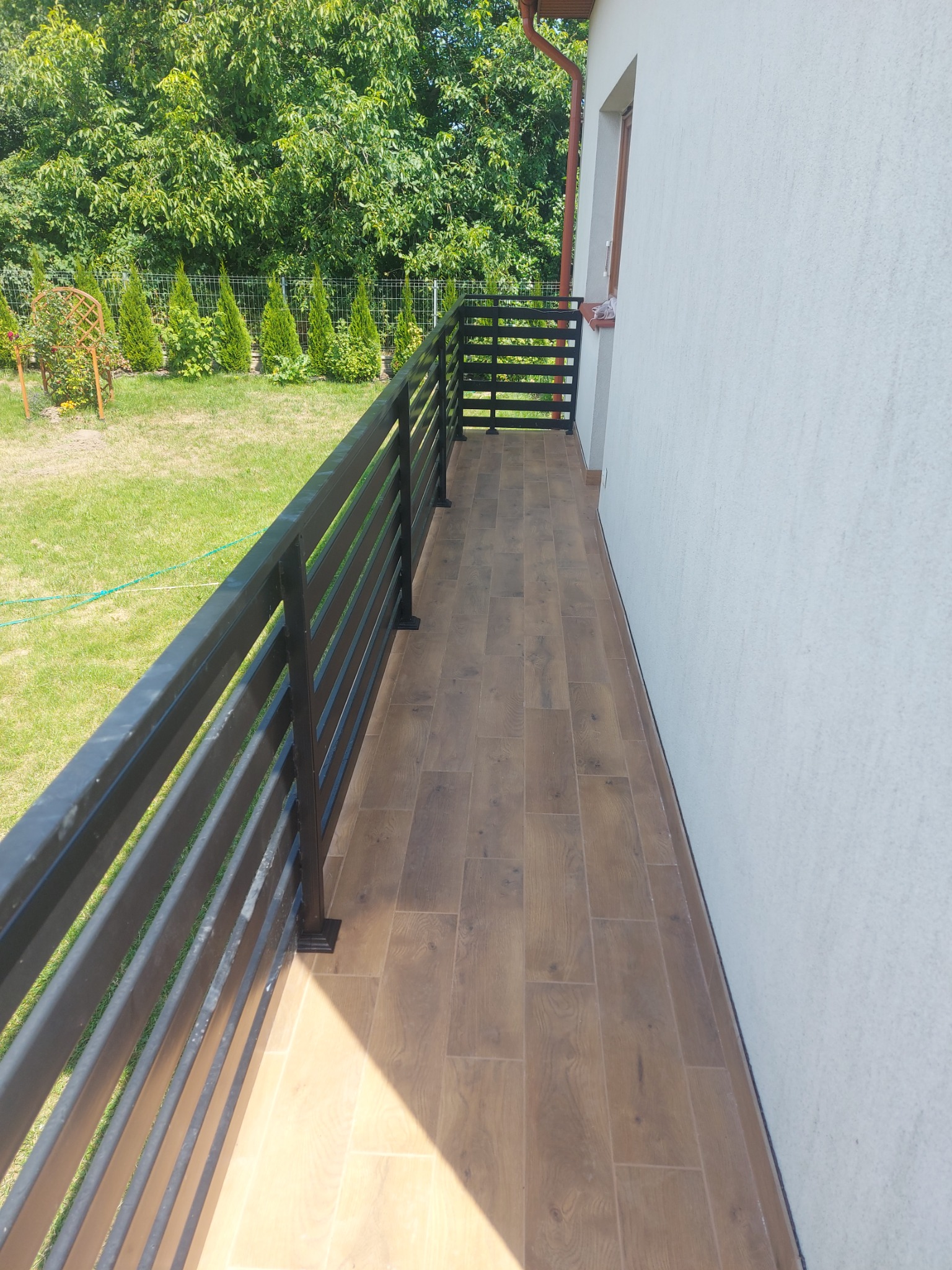 Wyłożony płytkami taras z czarną metalową balustradą, widok na trawnik i żywopłot z iglaków w słoneczny dzień.