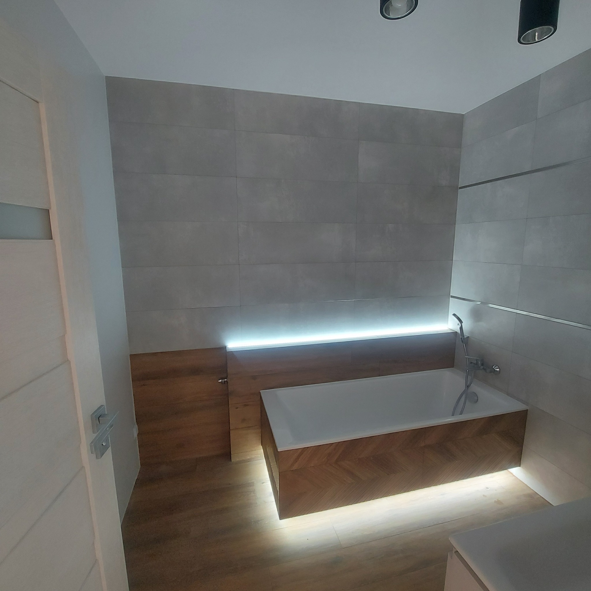 Nowoczesna łazienka z szarymi płytkami, podświetlaną wanną obudowaną drewnem w jodełkę i subtelnym oświetleniem punktowym.