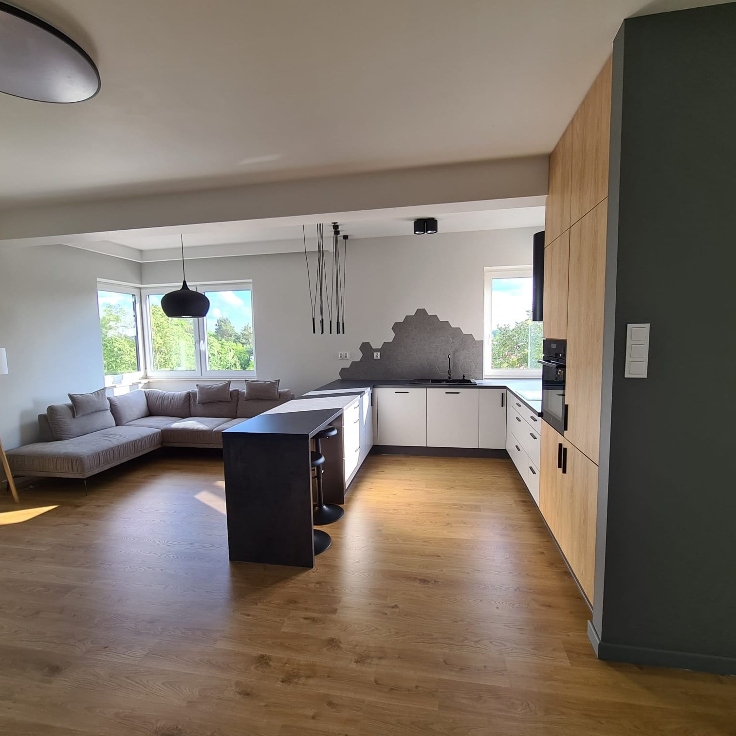 Nowoczesne wnętrze: salon z aneksem kuchennym, drewniana podłoga, szara sofa narożna, białe szafki kuchenne z czarnymi uchwytami, wyspa z hokerami, dekoracyjna ściana z szarych heksagonów...