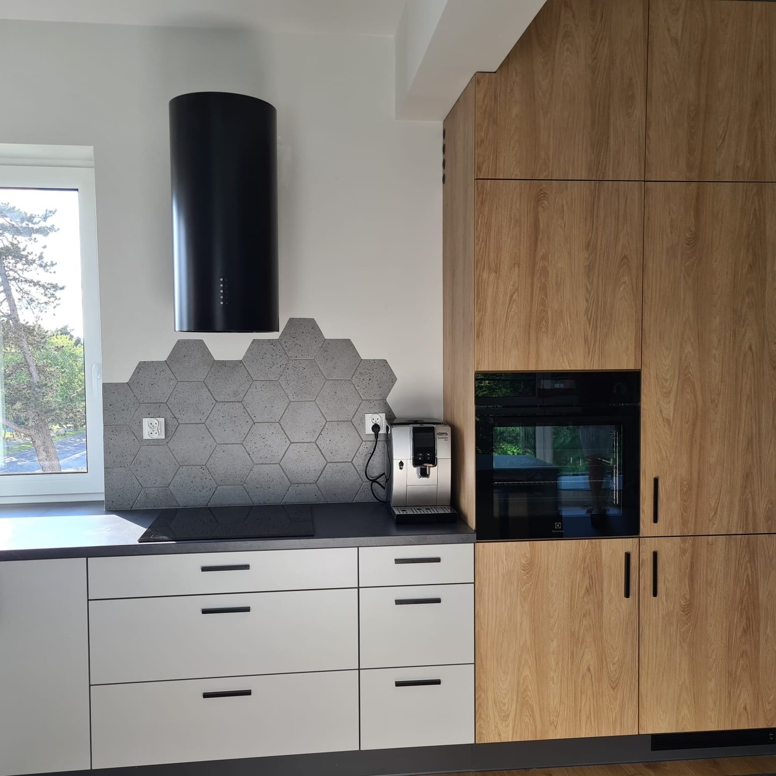 Nowoczesna kuchnia z drewnianymi szafkami, białymi szufladami, czarnym okapem cylindrycznym i szarą mozaiką heksagonalną nad blatem, z widocznym ekspresem do kawy.
