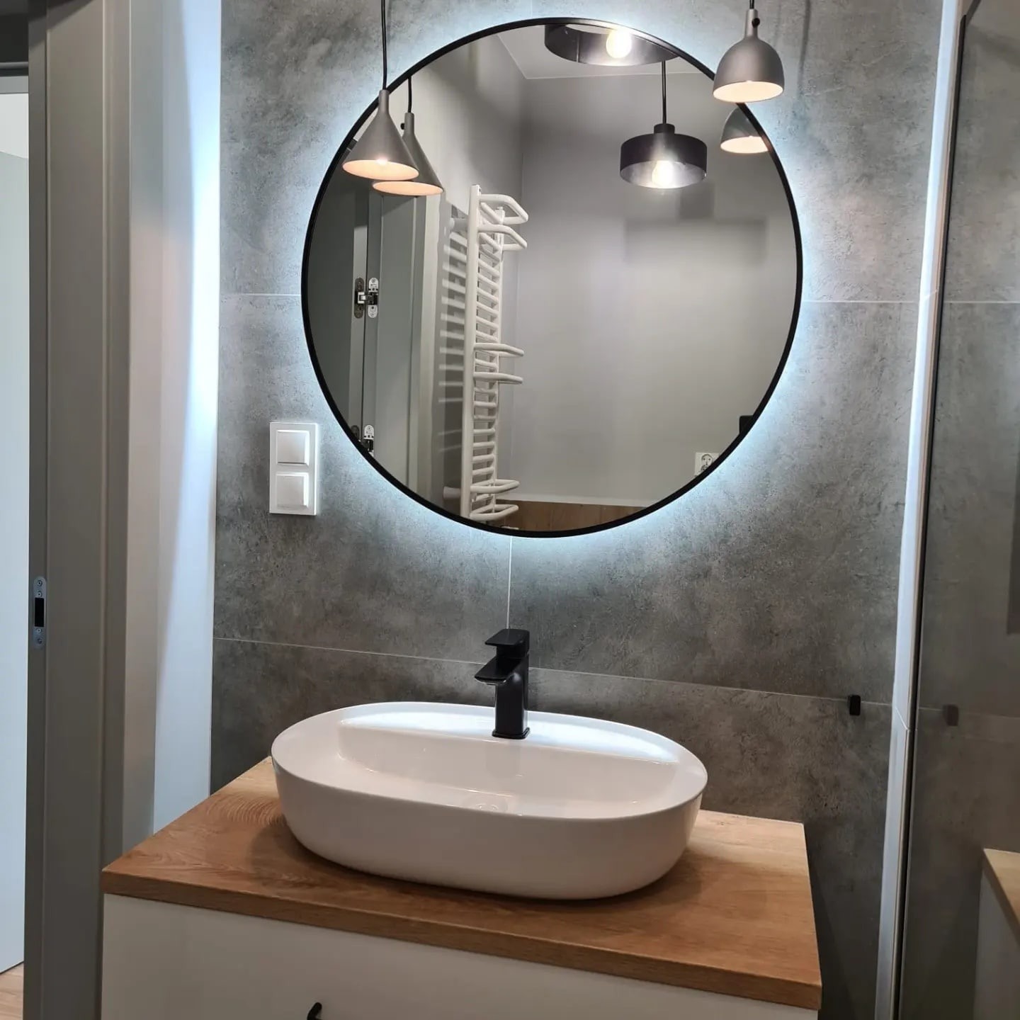 Nowoczesna łazienka z owalną umywalką nablatową, czarną armaturą i okrągłym lustrem z podświetleniem LED na tle szarych płytek imitujących beton.