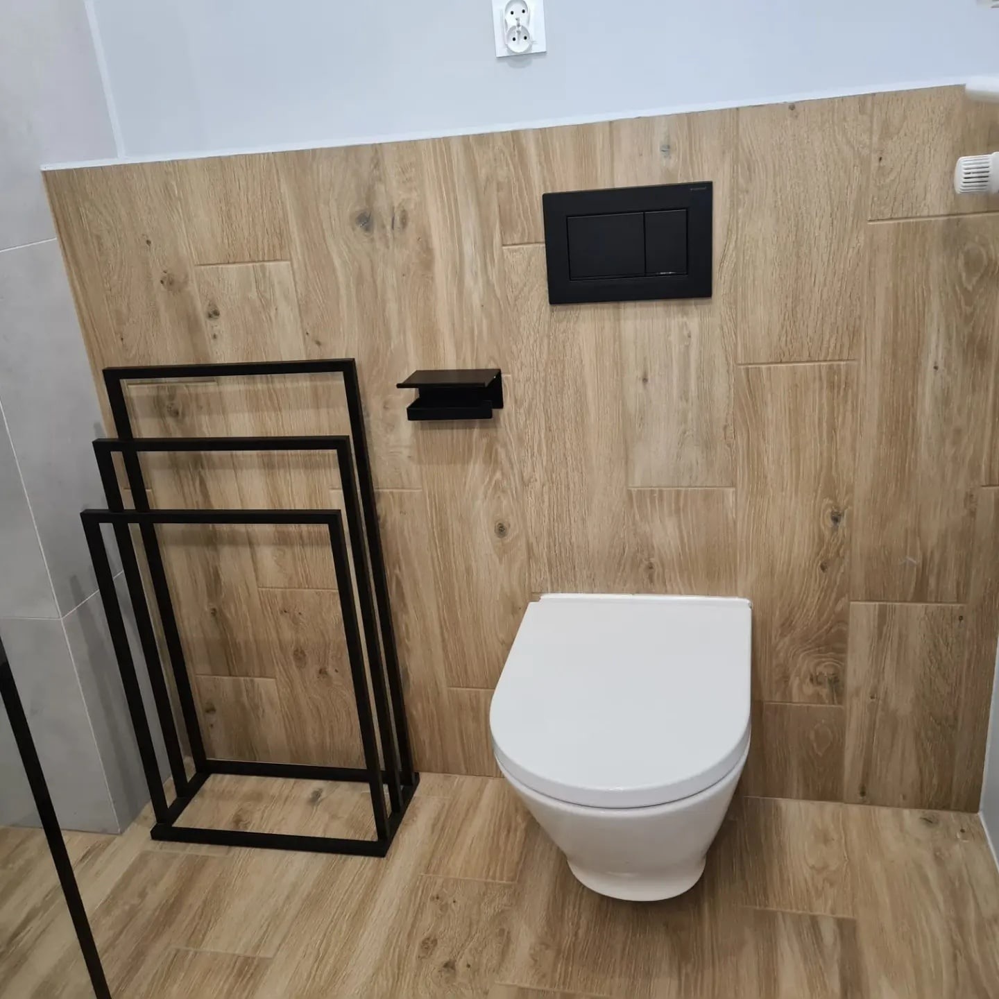 Nowoczesna łazienka z toaletą wiszącą, drewnopodobnymi płytkami na ścianach i podłodze, czarnym stelażem na ręczniki i czarnym przyciskiem spłukującym.
