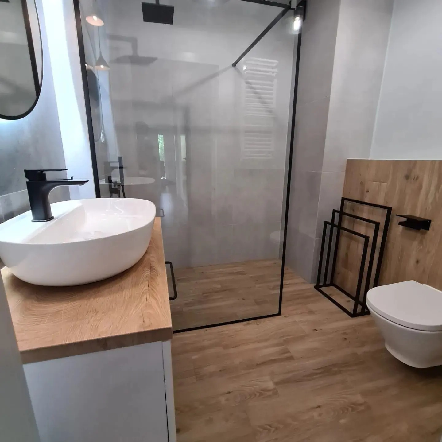 Nowoczesna łazienka z kabiną prysznicową typu walk-in, umywalką nablatową na drewnianym blacie, toaletą i grzejnikiem drabinkowym za szklaną ścianką.