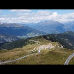 Spotlight Films Jakub Kiełtucki - Panoramiczny widok z lotu ptaka na krętą drogę górską, otoczoną zielonymi wzgórzami i szczytami. W tle błękitne niebo z chmurami. Widoczny czerwony samochód na serpentynach.