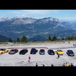 Spotlight Films Jakub Kiełtucki - Panoramiczne ujęcie rzędu luksusowych aut na parkingu z widokiem na góry. Osoby spacerują wokół pojazdów, tworząc dynamiczną scenę na tle malowniczego krajobrazu.