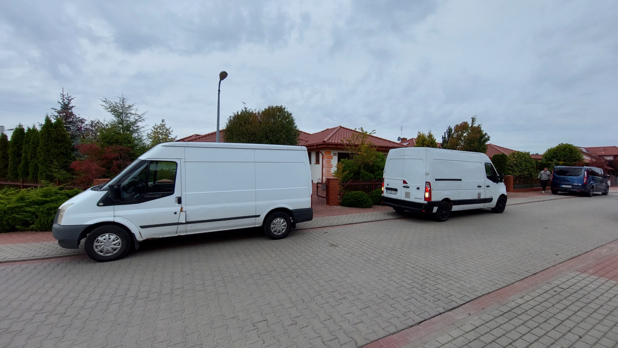 Dwa białe busy transportowe zaparkowane na ulicy przed domem jednorodzinnym, jeden z widocznym napisem 'Transit' na przedniej szybie.