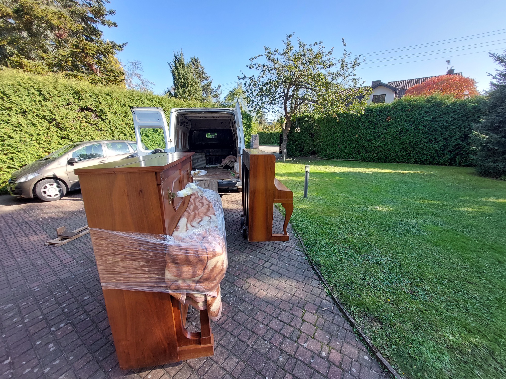 Dwa pianina, jedno owinięte folią i kocem, drugie bez zabezpieczeń, obok otwartego samochodu dostawczego, przygotowane do transportu na zewnątrz, na tle zielonego trawnika i żywopłotu.