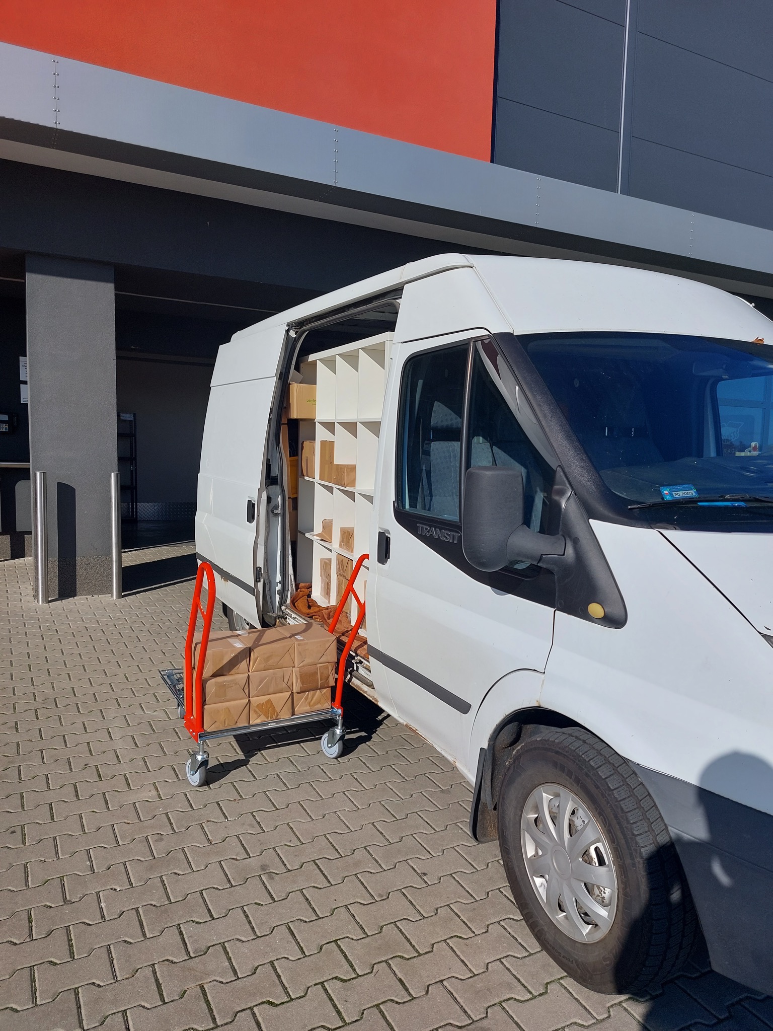 Biały furgon z otwartymi drzwiami, wypełniony kartonami i regałami, zaparkowany przed budynkiem z pomarańczowo-szarą fasadą; wózek transportowy z zapakowanymi przesyłkami obok pojazdu.