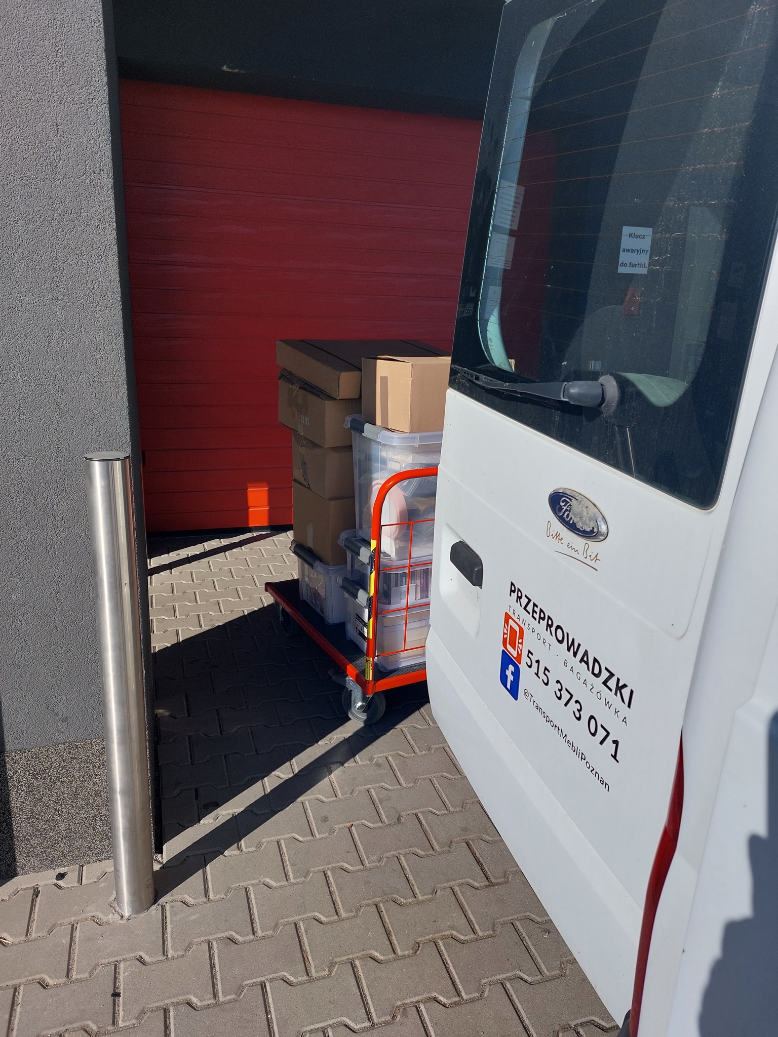 Wózek transportowy z kartonami i plastikowymi pojemnikami, częściowo widoczny z tyłu białego samochodu dostawczego z napisem 'Przeprowadzki' i numerem telefonu, zaparkowanego obok czerwonej bramy...