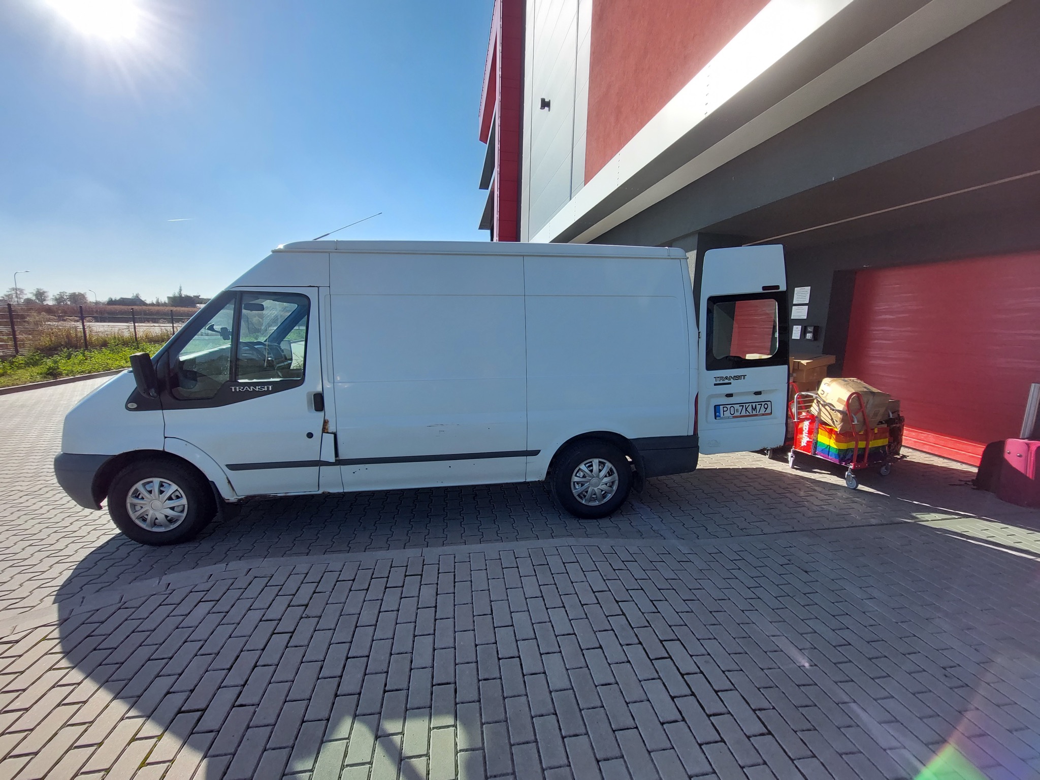 Biały samochód dostawczy Ford Transit z otwartymi tylnymi drzwiami, zaparkowany na brukowanej powierzchni przed czerwono-szarym budynkiem. W tle widoczny wózek transportowy z kartonami i tęczowymi...