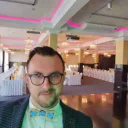 Mężczyzna w muszce na tle sali bankietowej z nakrytymi stołami, kryształowymi żyrandolami i różowym oświetleniem LED.