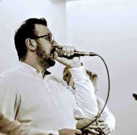 Wokalista z brodą i okularami śpiewa do mikrofonu podczas występu, w tle widoczni inni muzycy.