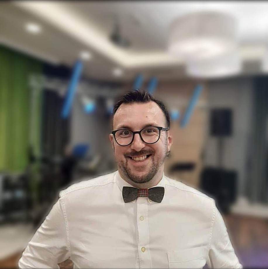 Portret uśmiechniętego mężczyzny w okularach i koszuli z muchą na tle rozmytego wnętrza sali z oświetleniem eventowym.