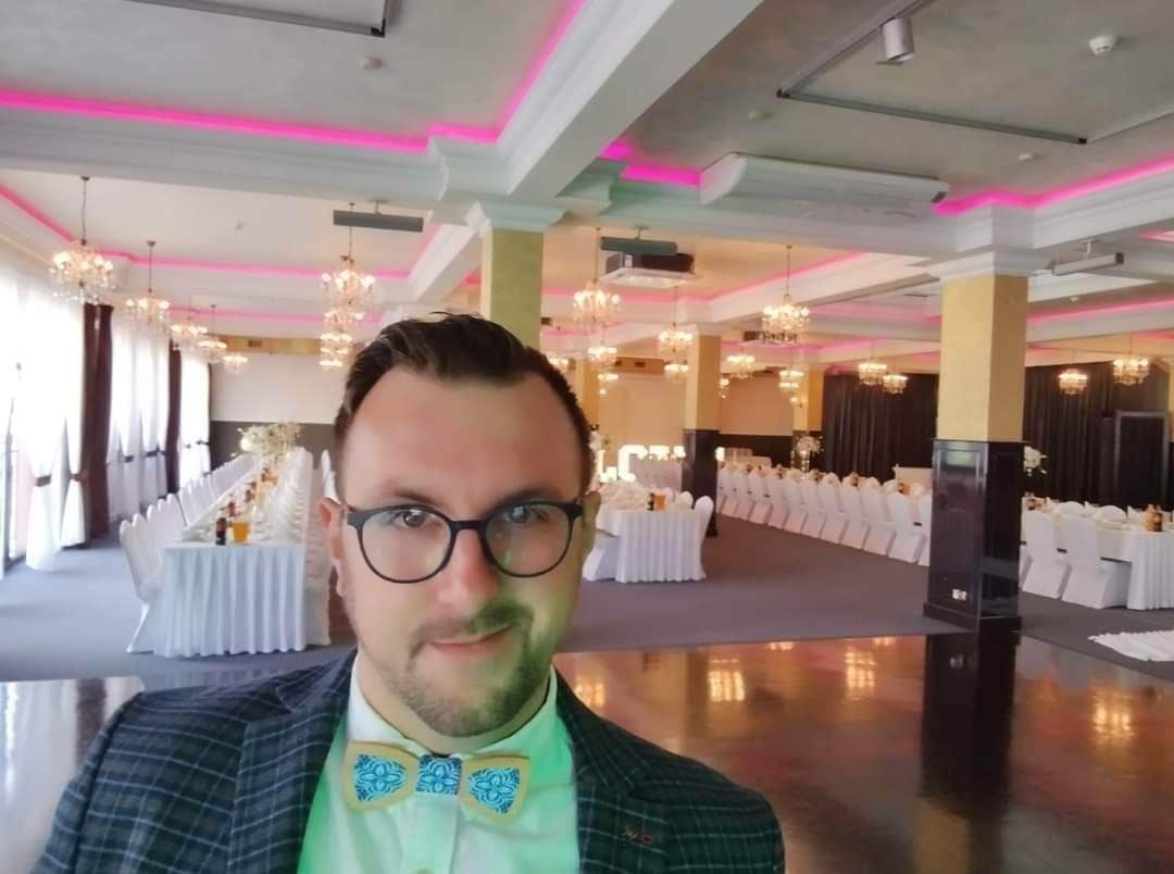 Mężczyzna w muszce na tle sali bankietowej z nakrytymi stołami, kryształowymi żyrandolami i różowym oświetleniem LED.