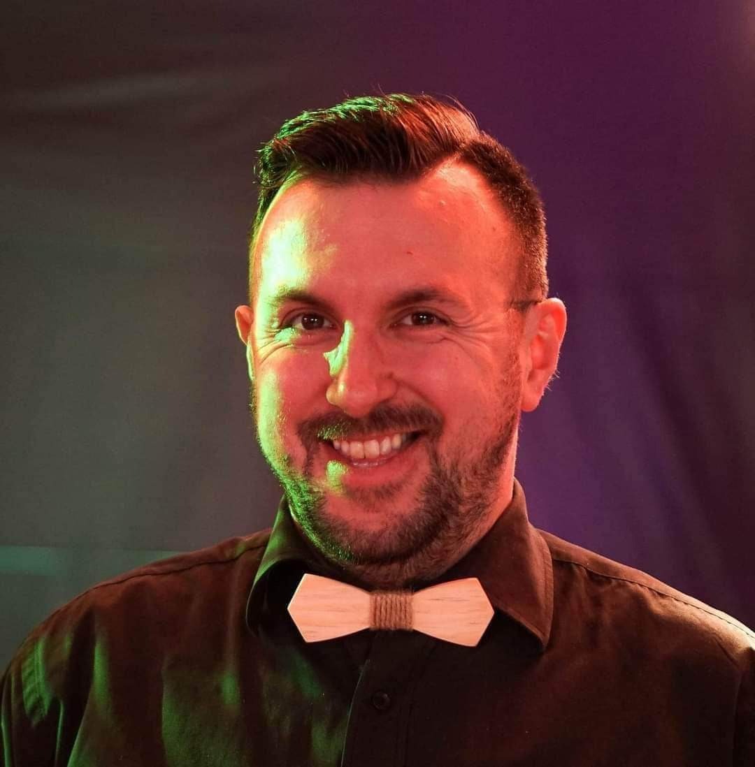 Uśmiechnięty mężczyzna z brodą w czarnej koszuli i drewnianej muszce, oświetlony zielonym i czerwonym światłem.