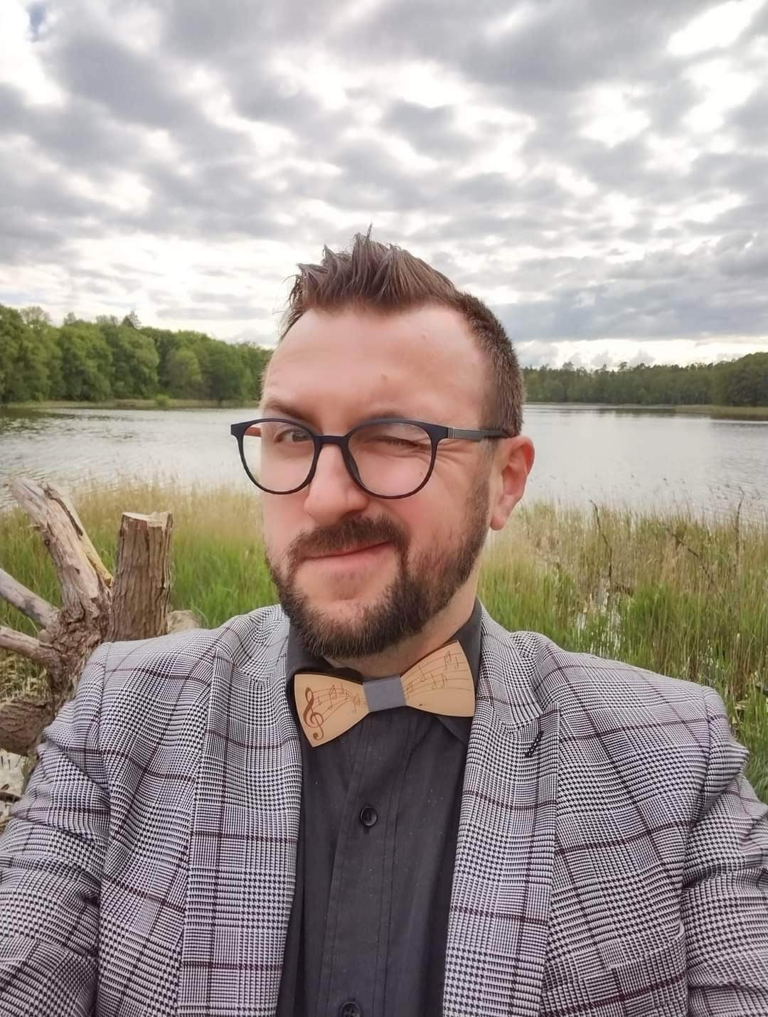 Mężczyzna w okularach mruga okiem, ubrany w marynarkę w kratę i drewnianą muszkę z motywem nut, na tle jeziora i drzew.