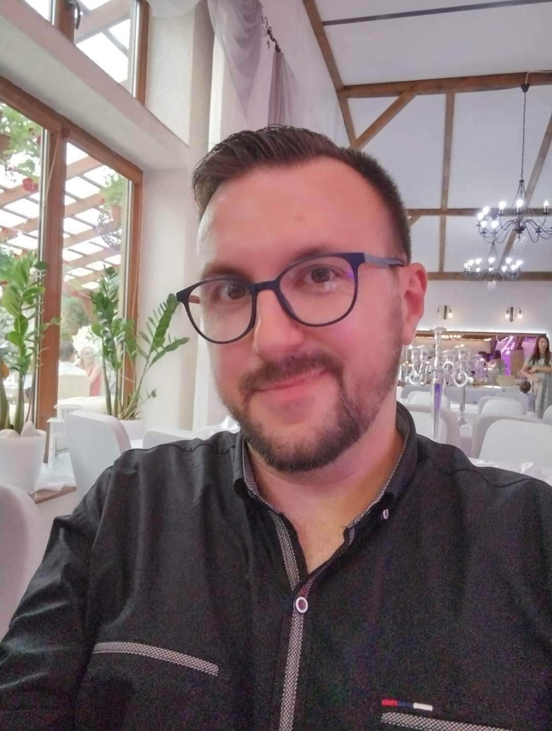 Mężczyzna w okularach i czarnej koszuli na tle sali bankietowej z dekoracjami i oświetleniem, widoczne stoły z białymi obrusami i krzesłami.
