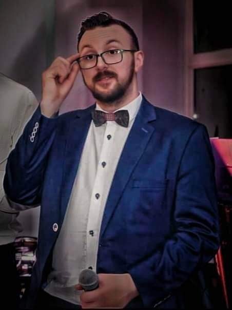 Mężczyzna w niebieskiej marynarce i muszce w kratę, poprawiający okulary, trzymający mikrofon w dłoni.