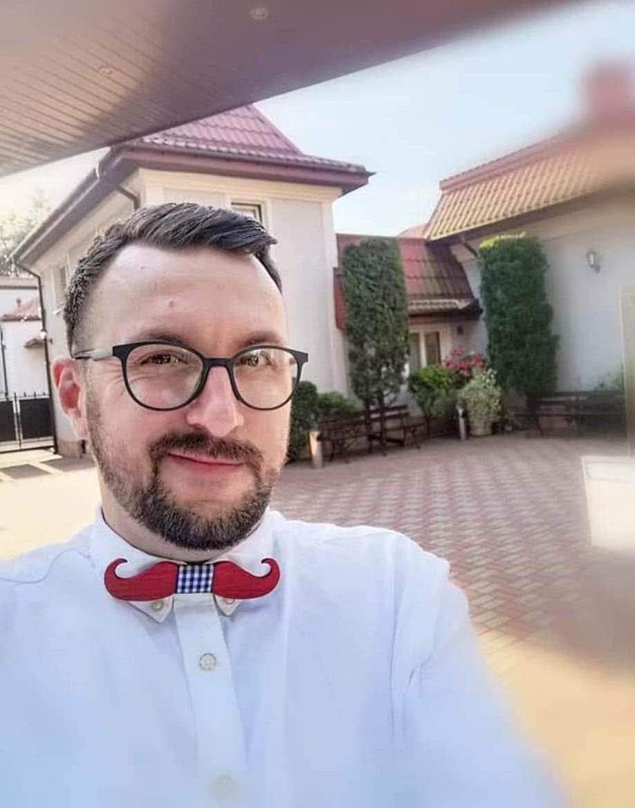 Mężczyzna w okularach i białej koszuli z muszką w kształcie wąsów na tle budynku z czerwoną dachówką i zadbanym ogrodem.