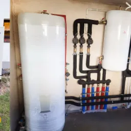 Biała pompa ciepła Panasonic na zewnątrz budynku, obok zbiornik akumulacyjny owinięty folią, oraz wiszący piec gazowy z podłączonymi rurami wewnątrz pomieszczenia w surowym stanie.