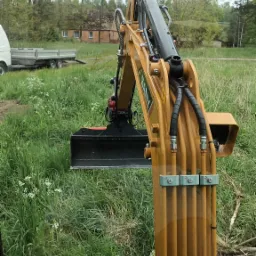 Żółta koparka SANY z czarną łyżką na zarośniętym terenie, w tle przyczepa i budynek z czerwonej cegły pod pochmurnym niebem.