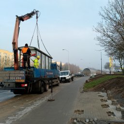 Marbruk - Montaż przystanku autobusowego z użyciem dźwigu HDS na tle miejskiej ulicy i częściowo ułożonej kostki brukowej. Widoczni pracownicy w odblaskowych ubraniach.