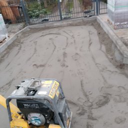Marbruk - Wyrównywanie piasku pod kostkę brukową za pomocą zagęszczarki Wacker Neuson, widoczne ślady po przejazdach. Przygotowany teren ograniczony obrzeżami.