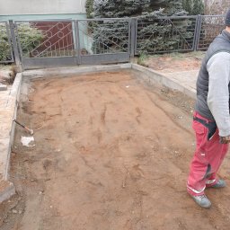 Marbruk - Przygotowany teren pod kostkę brukową z obrzeżami, widoczna łopata i fragment ogrodzenia. Pracownik w czerwonych spodniach w tle. Etap prac ziemnych.