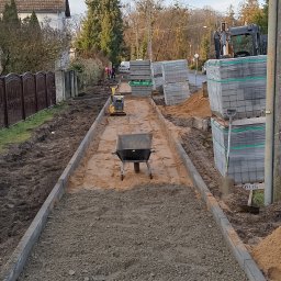 Marbruk - Przygotowanie podłoża pod kostkę brukową w Gorzowie Wielkopolskim: wyrównany teren, obrzeża, piasek, ubijarka i taczka. Widoczne palety z kostką i pracownicy w tle.