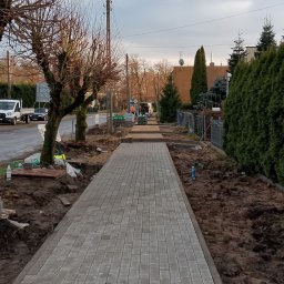 Marbruk - Świeżo ułożony chodnik z szarej kostki brukowej wzdłuż ulicy, z widocznymi elementami przygotowania terenu i pracownikami w oddali. Uwagę zwraca regularny wzór kostki.