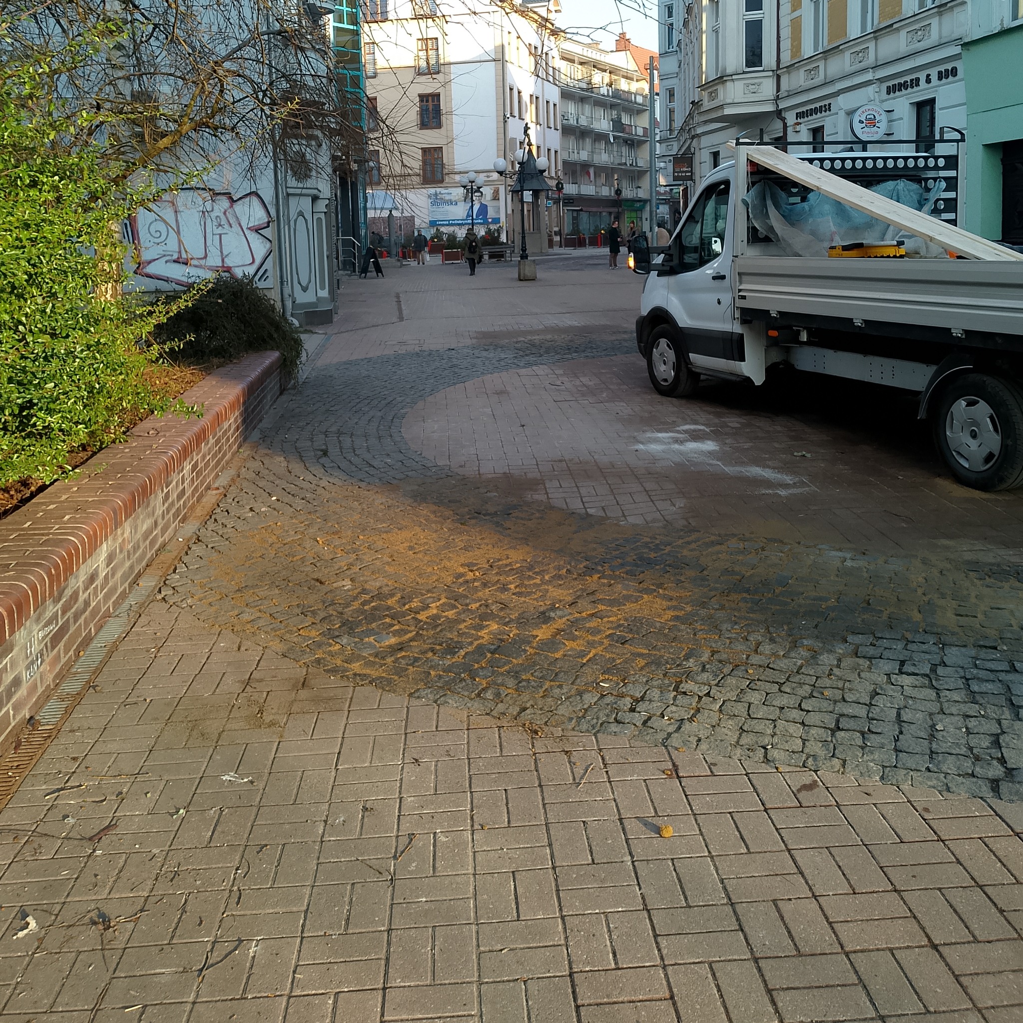 Ulica wyłożona kostką brukową i granitową, z widocznym samochodem dostawczym w tle. Fragment chodnika z rozsypanym piaskiem. Widok na budynki w tle.