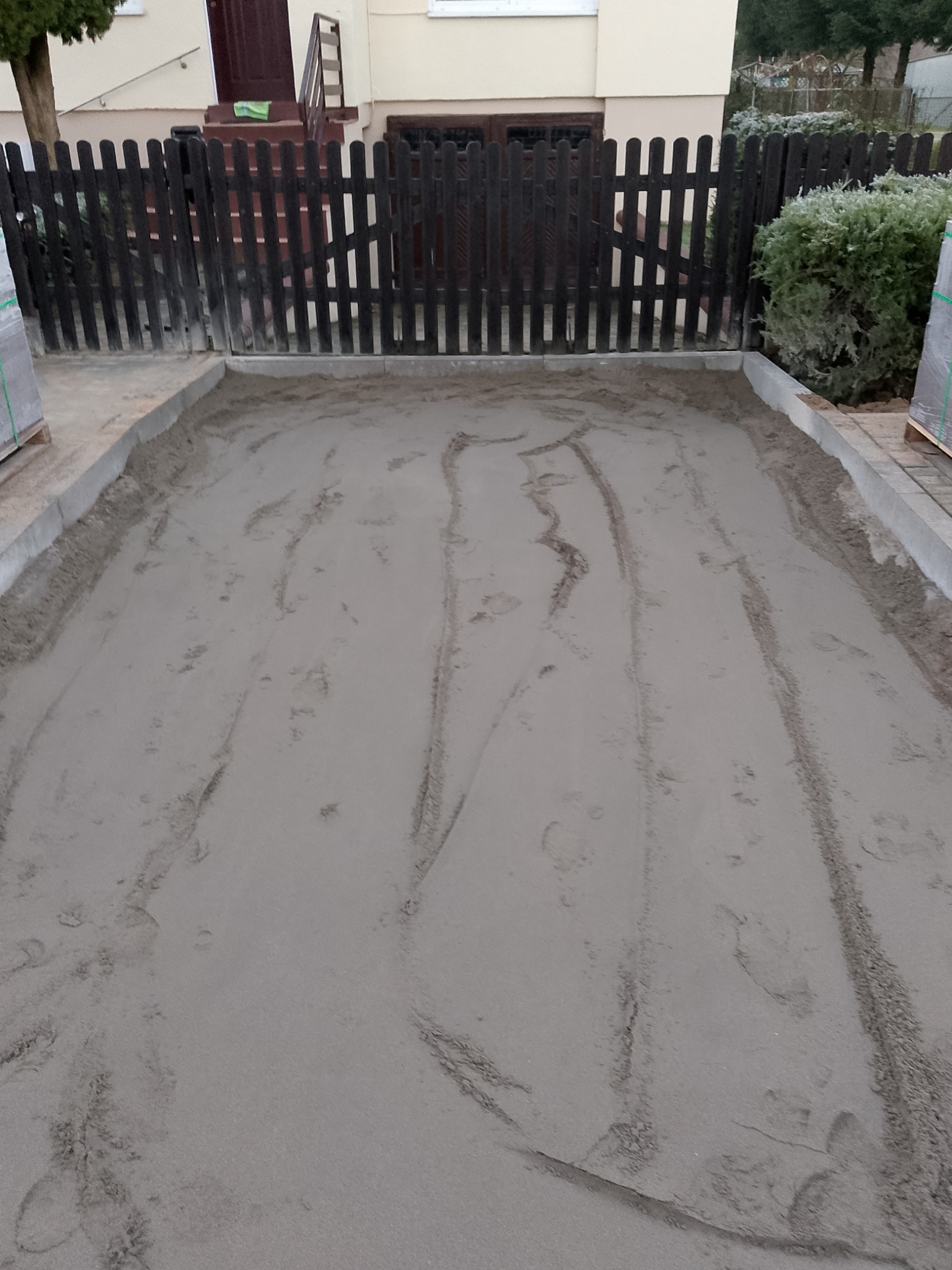 Przygotowany teren pod kostkę brukową z widocznymi śladami stóp, obrzeżami i paletą kostki w tle. Widok na posesję z drewnianym ogrodzeniem.