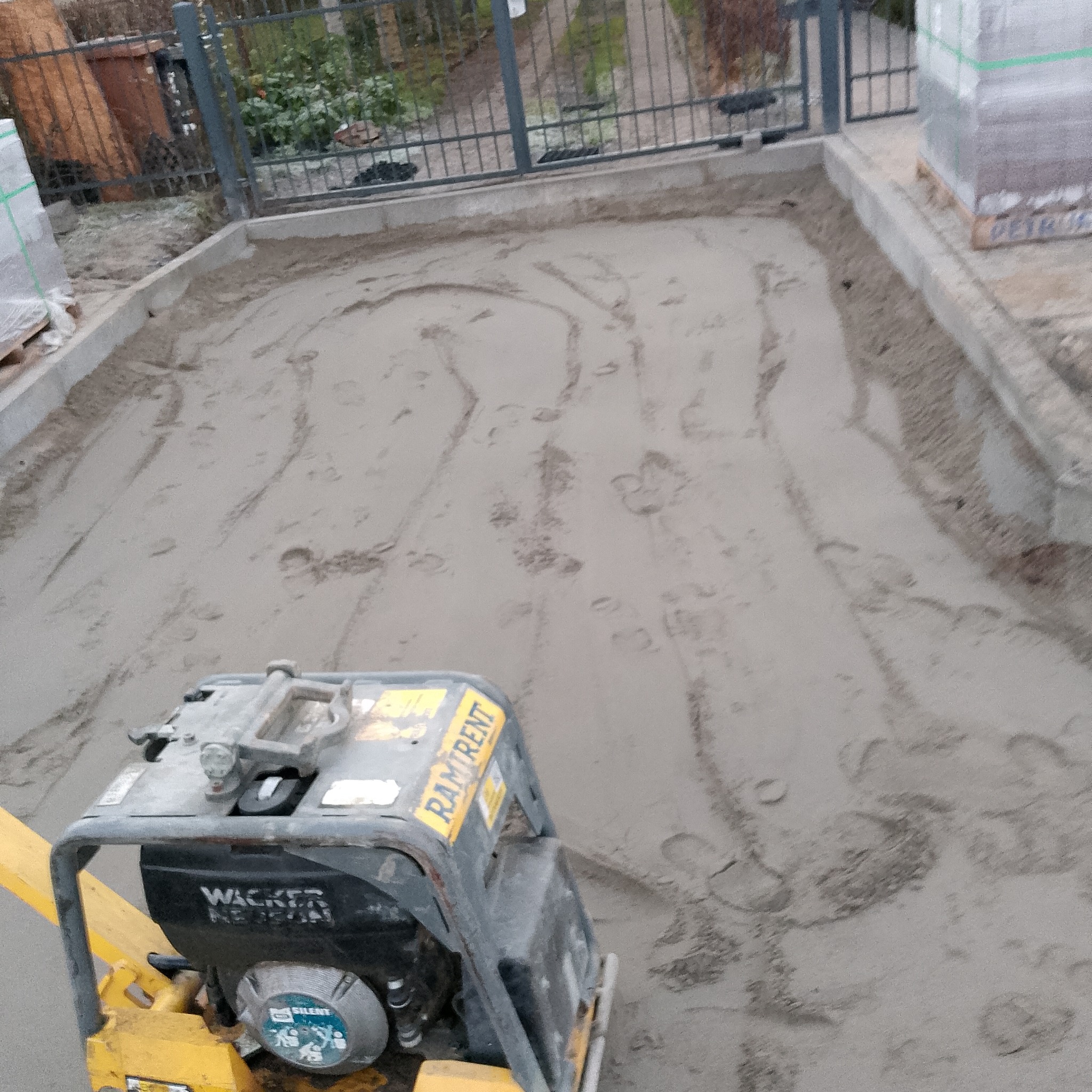 Wyrównywanie piasku pod kostkę brukową za pomocą zagęszczarki Wacker Neuson, widoczne ślady po przejazdach. Przygotowany teren ograniczony obrzeżami.